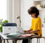 Femme travaillant de chez elle avec une imprimante couleur Xerox C310, fournie par D&O Partners, illustrant une intégration harmonieuse dans un espace de travail à domicile.