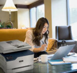 Een vrouw werkt vanuit huis aan haar laptop met een Xerox C235 multifunctionele kleurenprinter op de achtergrond, wat een flexibele thuiswerkoplossing toont.