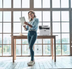 A smiling woman standing in a bright, spacious office reading a document next to a Xerox B235 multifunction printer, illustrating the efficiency and convenience of Xerox printing solutions supplied by D&O Partners.