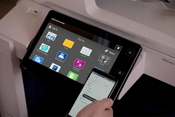 Gros plan de l'écran tactile interactif ConnectKey sur une imprimante Xerox AltaLink C8045 avec une personne utilisant un smartphone via l'impression mobile. L'écran affiche diverses icônes d'application pour faciliter l'utilisation, illustrant la façon dont la technologie favorise l'intégration et la convivialité dans le bureau moderne.