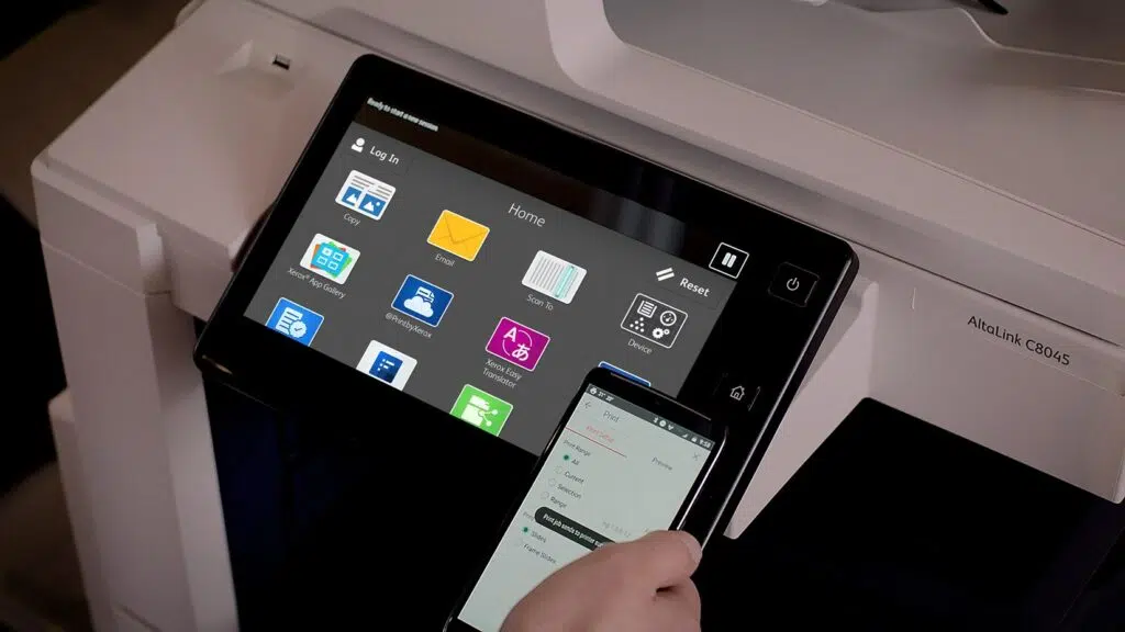 Close-up van het ConnectKey interactieve touchscreen op een Xerox AltaLink C8045 printer met een persoon die een smartphone bedient via mobiel printen. Het scherm toont diverse applicatie-iconen voor gebruiksgemak, illustrerend hoe technologie integratie en gebruiksvriendelijkheid in het moderne kantoor bevordert.