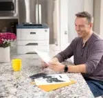 Een man glimlachend terwijl hij een vers geprint document bekijkt van de Xerox B310 printer in een gezellige thuisomgeving met een marmeren keukenaanrecht, een boeket bloemen en een smartphone op de achtergrond. Dit tafereel weerspiegelt het gemak en het gebruiksgemak van de printer in een dagelijkse, huiselijke setting.