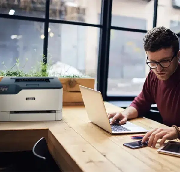 Imprimante couleur Xerox C230 de D&O Partners à côté d'un homme travaillant sur son portable dans un espace de coworking.