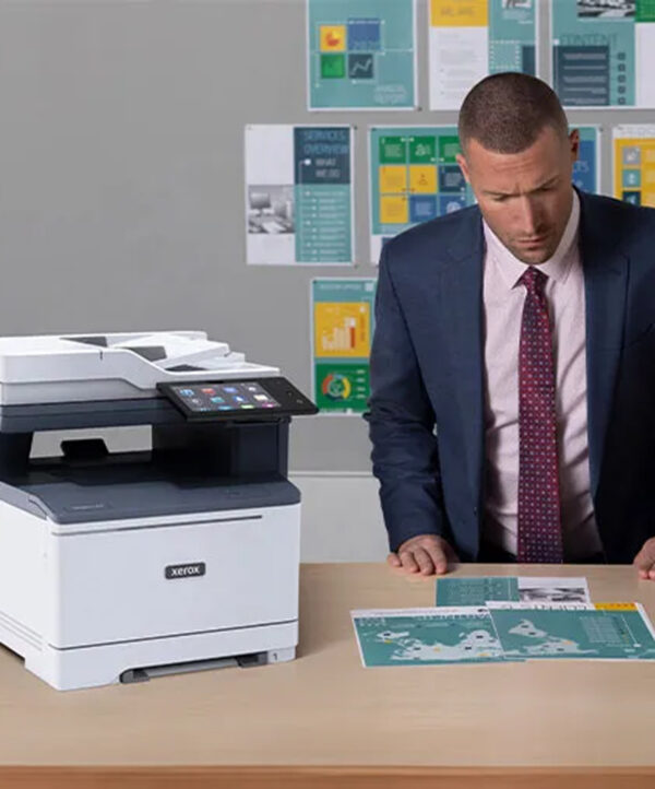 Serious businessman reviewing documents in front of a Xerox VersaLink C415 printer in a professional setting with company information posters in the background.