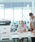 D&O Partners' Xerox VersaLink C625 printer installed next to two professionals talking in a bright, modern office, illustrating a collaborative working environment.