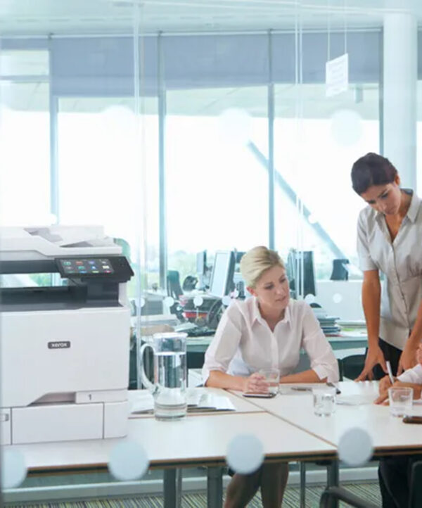 Imprimante Xerox VersaLink C625 de D&O Partners installée à côté de deux professionnels discutant dans un bureau lumineux et moderne, illustrant une scène de travail collaborative.