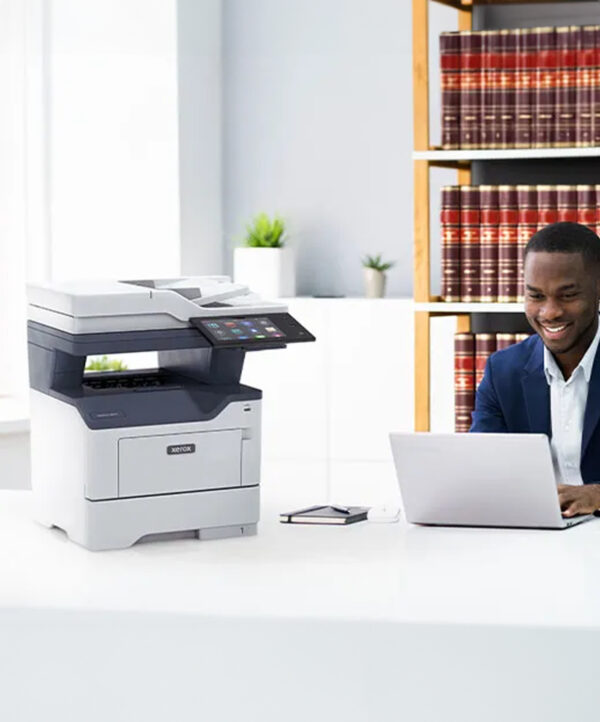 Moderne kantooromgeving met een Xerox VersaLink B415 multifunctionele printer op de voorgrond en een vrolijke zakenman die op een laptop werkt aan een bureau op de achtergrond. Daarnaast beschikt de printer over een aanraakscherminterface.