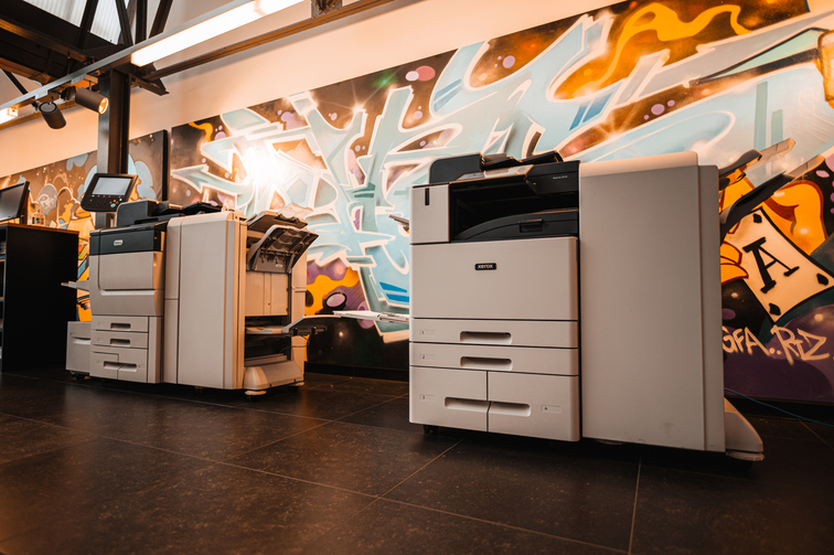 Twee Xerox kantoorprinters in de showroom van D&O Partners, de Xerox concessie in Brussel. Links staat de Xerox PrimeLink C9065 en rechts de Xerox AltaLink C8155, beide zien er robuust en professioneel uit, klaar voor gebruik in een dynamische werkomgeving.