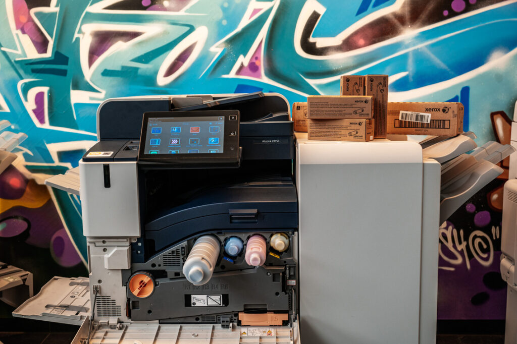Gros plan sur une imprimante multifonction Xerox AltaLink C8155 avec un écran tactile interactif et une sélection de consommables sur le dessus. L'accent est mis sur les détails technologiques et les fournitures essentielles au travail de bureau quotidien.